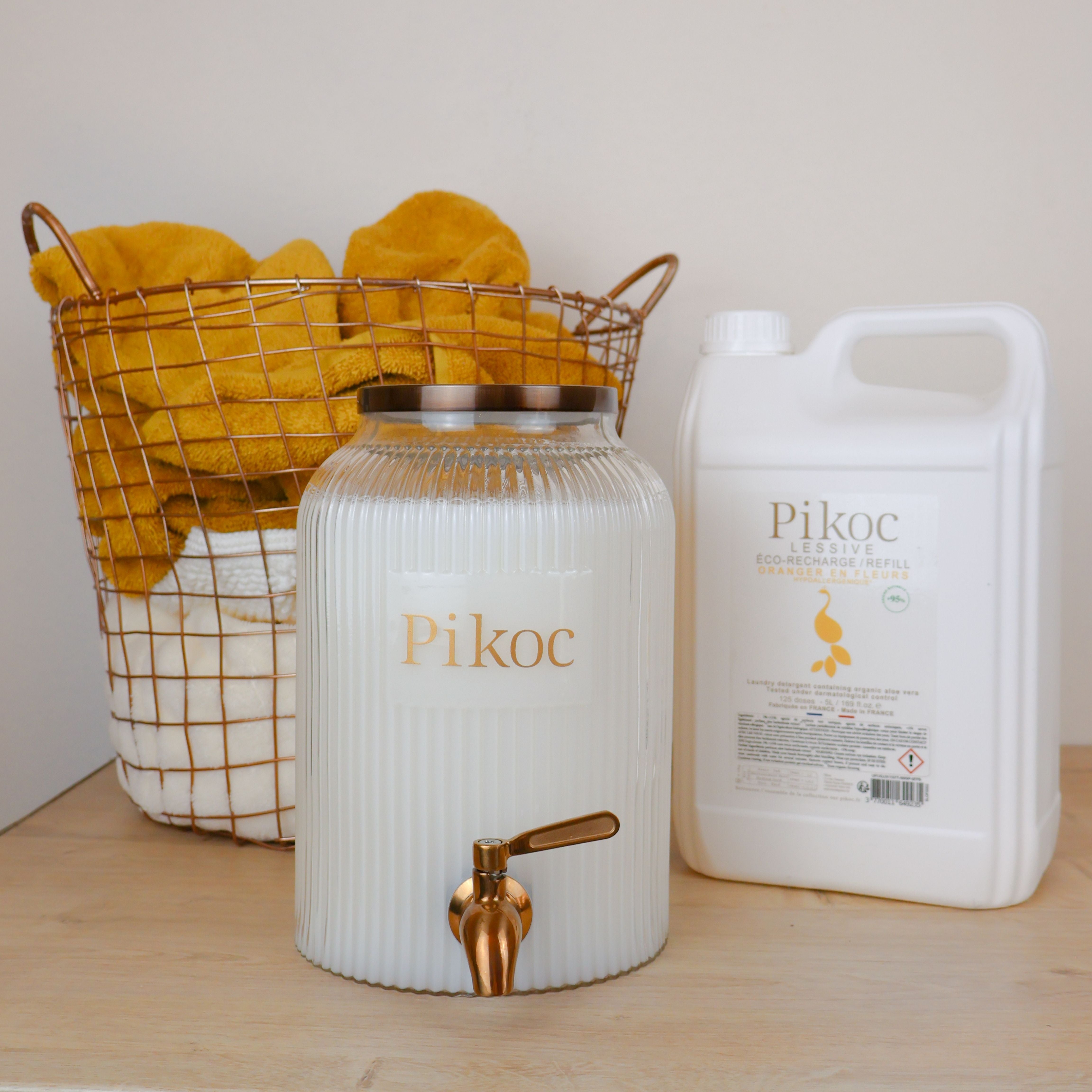 Fontaine à lessive 4L et Éco-recharge 5L Pikoc, Oranger en Fleurs, posé sur un meuble en bois avec panier de linge