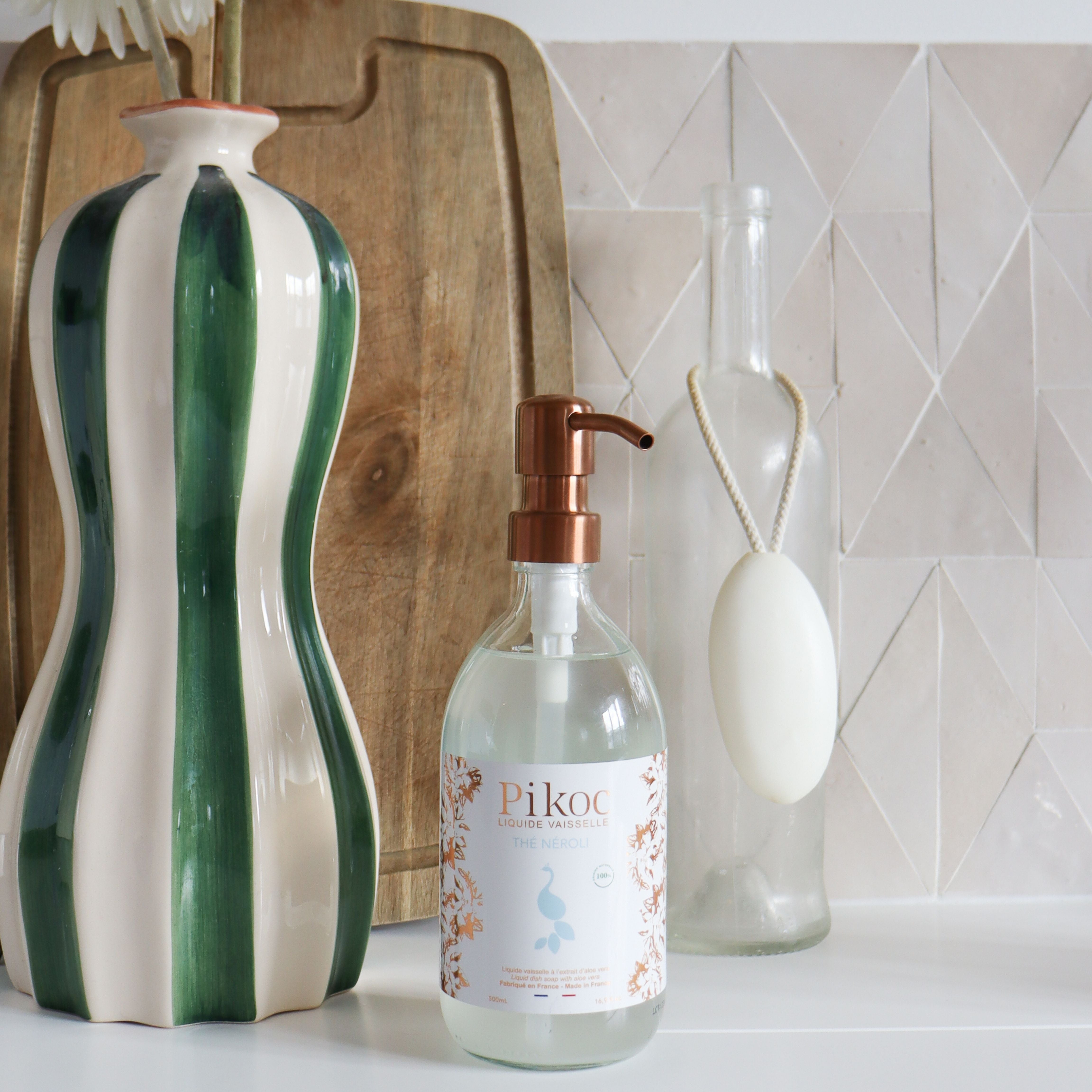 Liquide vaisselle Pikoc Thé Néroli, 490mL, posé à côté d’un vase, d’une bouteille d’eau et d’une planche à découper, ambiance cuisine