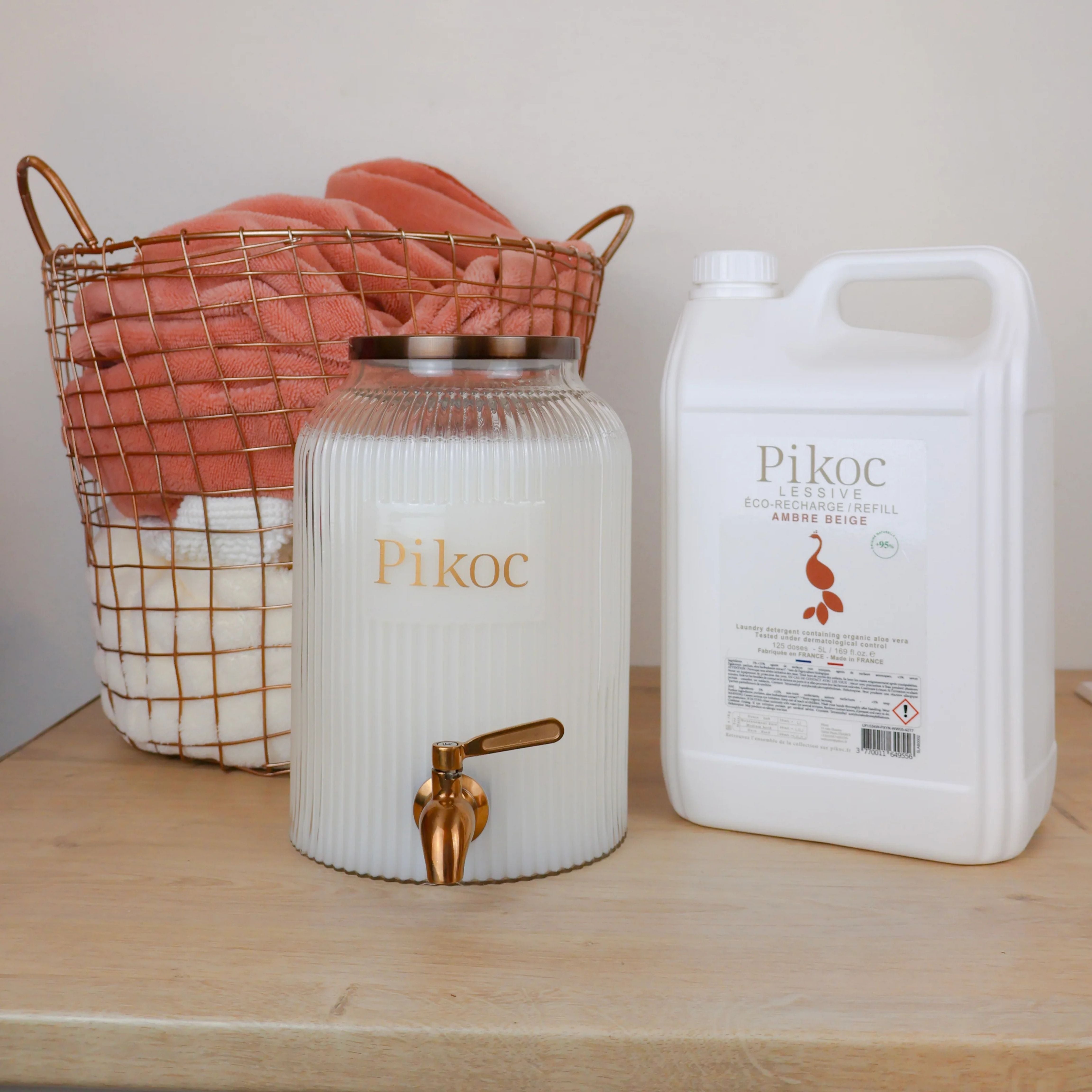 Fontaine à lessive 4L et Éco-recharge 5L Pikoc, Ambre Beige, posé sur un meuble en bois avec panier de linge