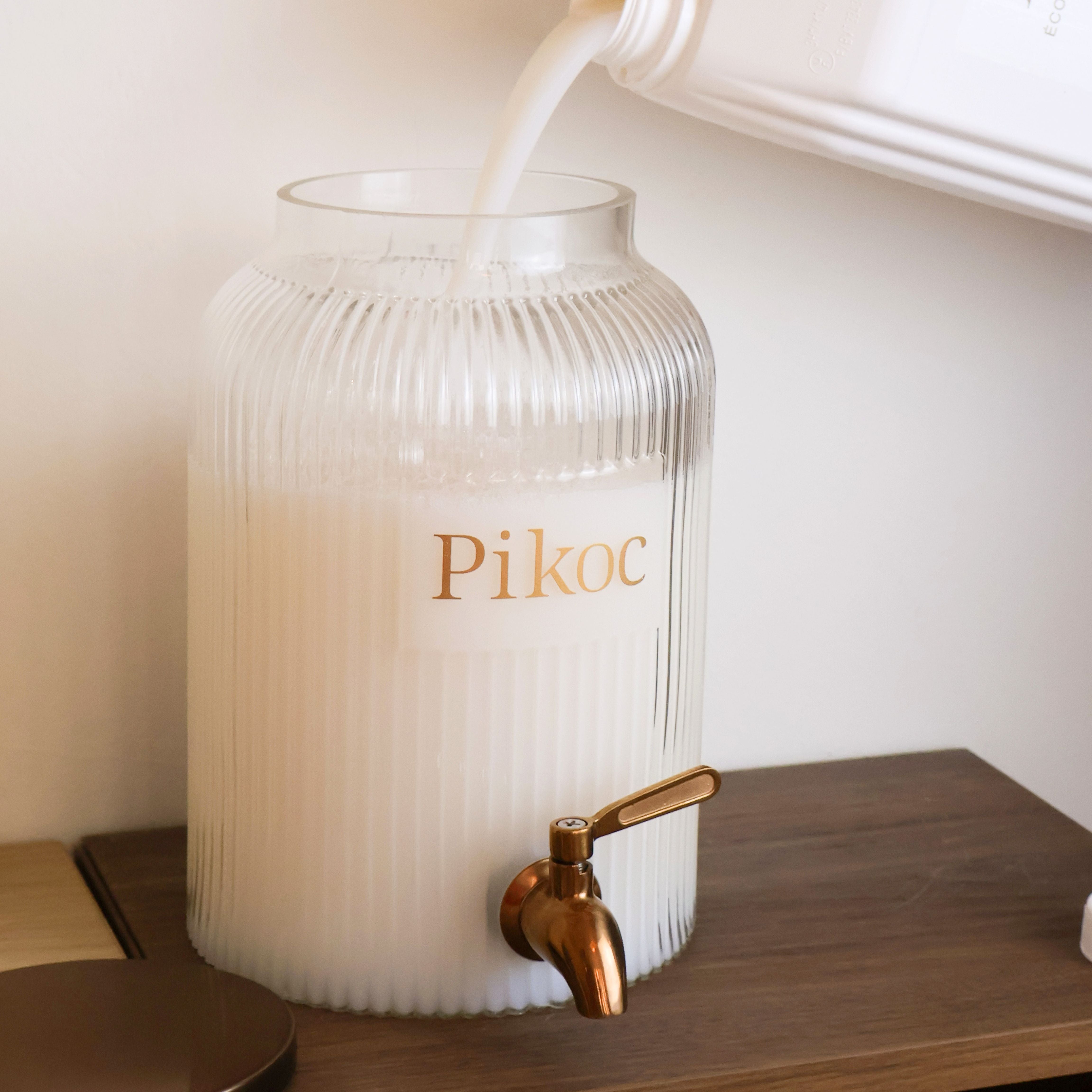 Fontaine à lessive Pikoc 4L, posé sur un meuble en bois, remplissage avec l’Éco-recharge de 5L de lessive, ambiance préparation